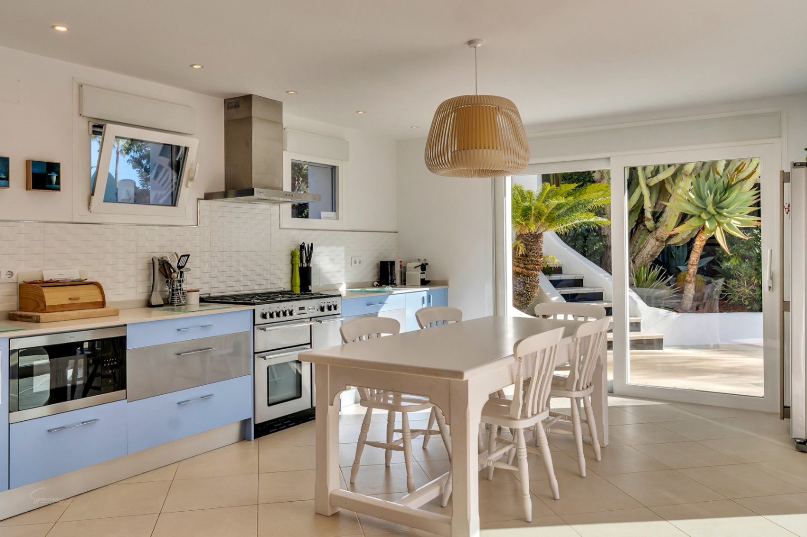 Mediterrane keuken met lichtblauwe kasten, een roestvrijstalen fornuis en een witte eethoek. Grote glazen schuifdeuren zorgen voor een directe verbinding met de zonnige tuin.