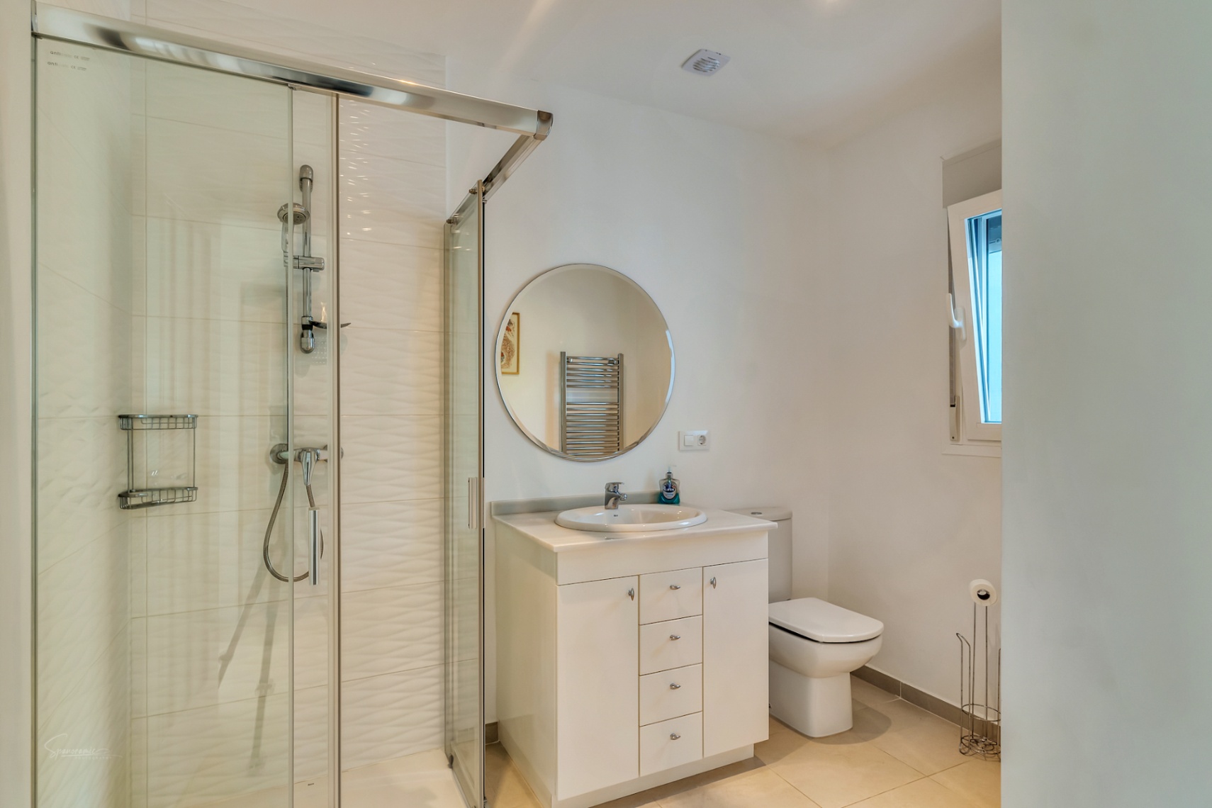 Moderne badkamer met glazen douchecabine, getextureerde tegels, wit wastafelmeubel met ronde spiegel en natuurlijk lichtinval via een zijraam.