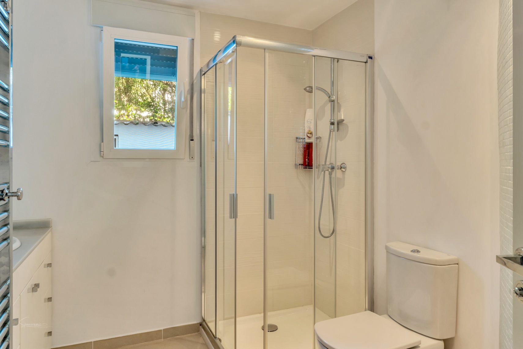 Moderne badkamer met glazen hoekdouche, wit wastafelmeubel met lades en een raam dat zorgt voor natuurlijk licht en uitzicht op de tuin.