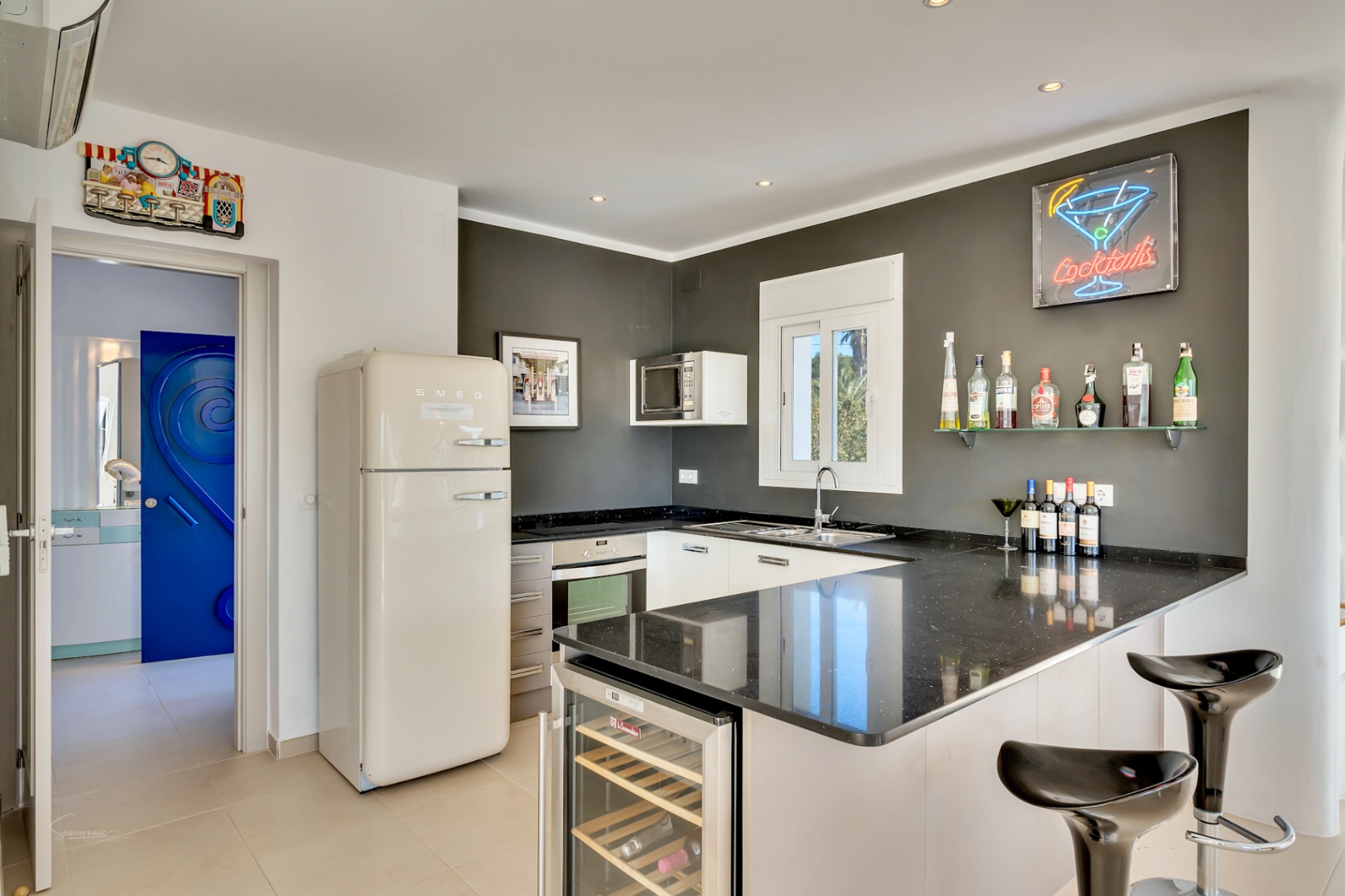 Moderne keuken met zwart granieten aanrechtblad, retro Smeg koelkast en een bar met geïntegreerde wijnkoeler en neonverlichting aan de grijze accentmuur.