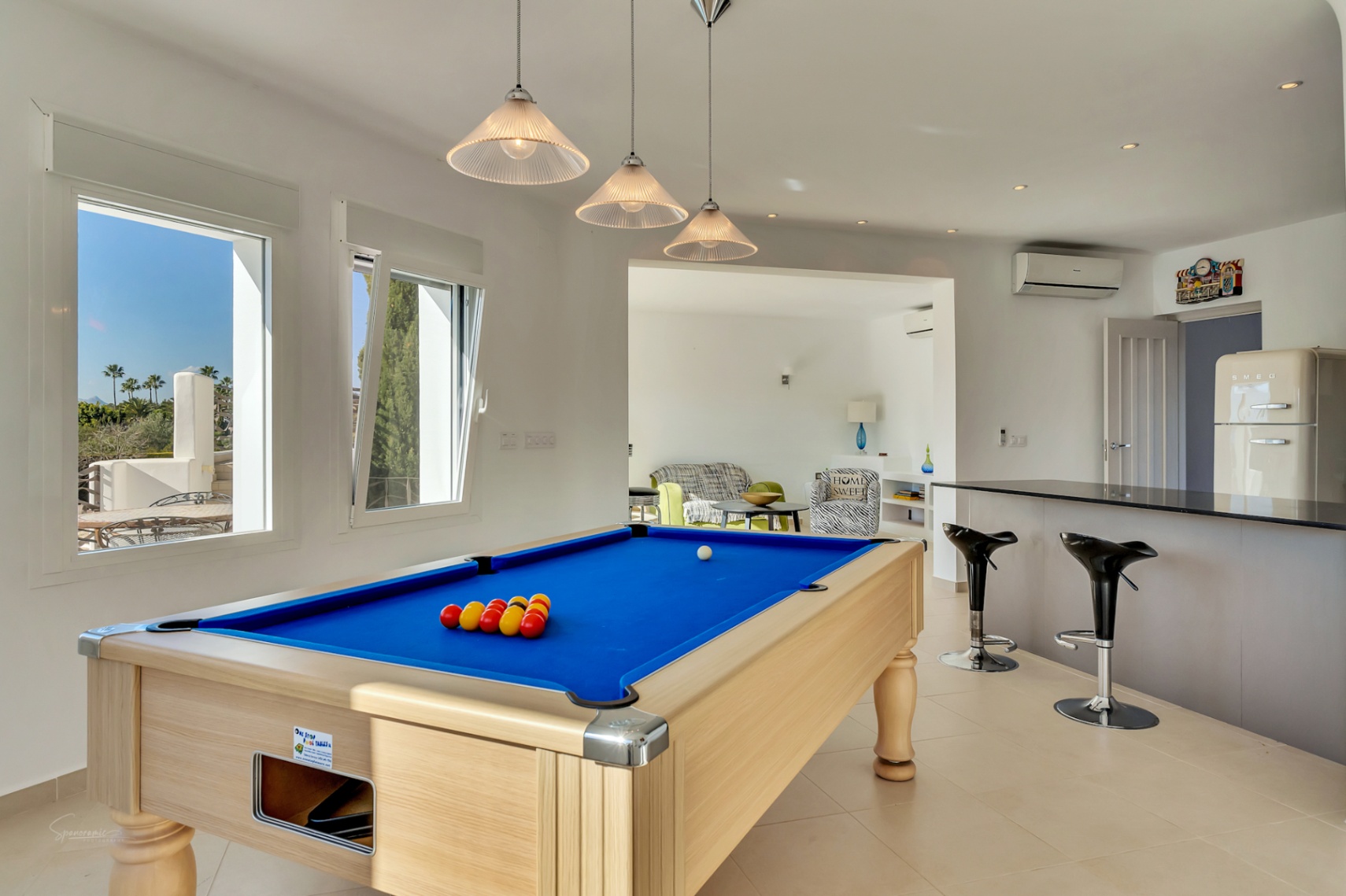 Ontspanningsruimte met een professionele biljarttafel, een moderne bar met krukken en een retro koelkast. Grote ramen bieden veel natuurlijk licht en zicht op de omgeving.
