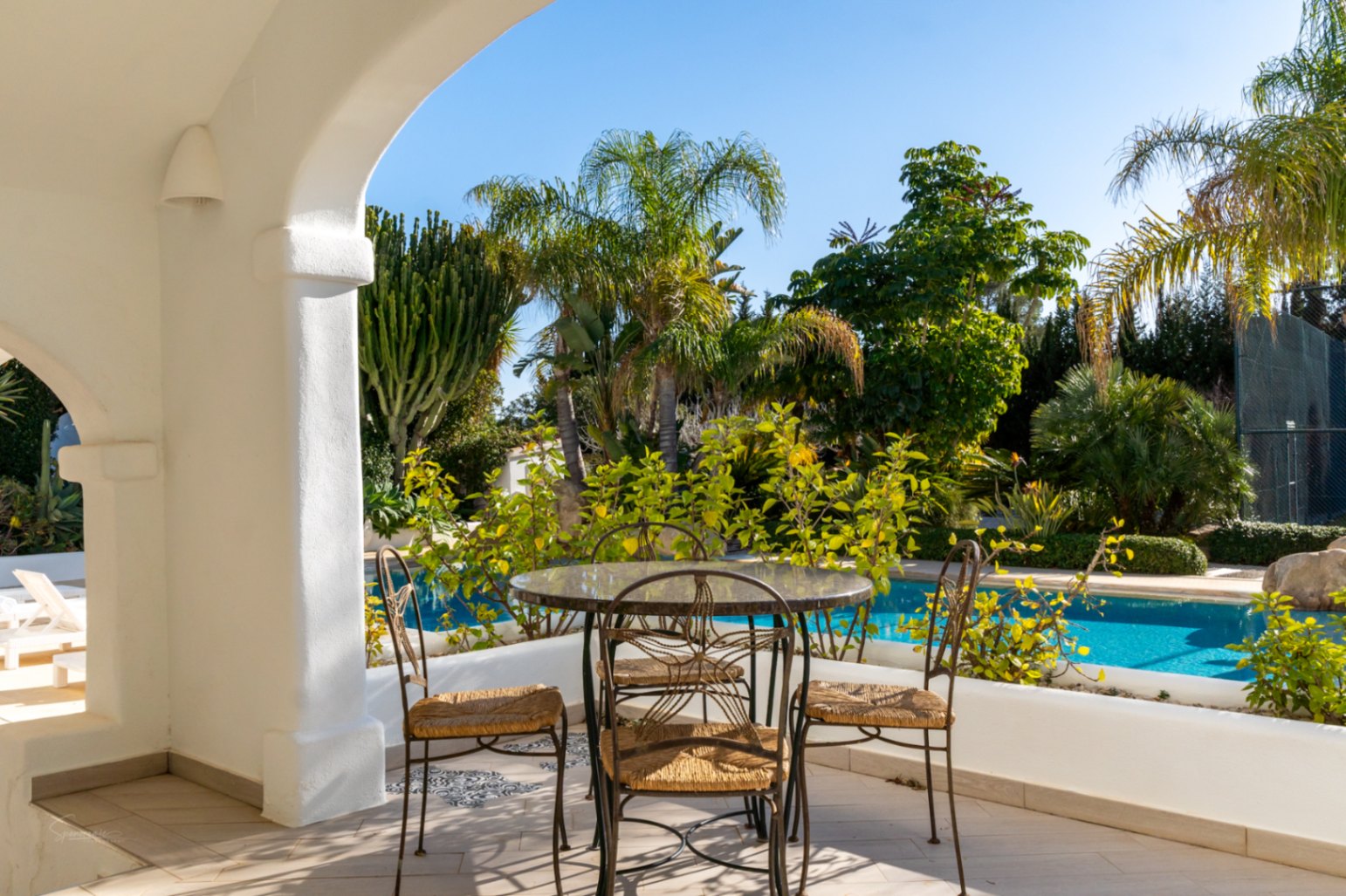 Overdekte naya met witte bogen en smeedijzeren meubilair, uitkijkend op het zwembad en de mediterrane tuin met palmbomen en cactussen.