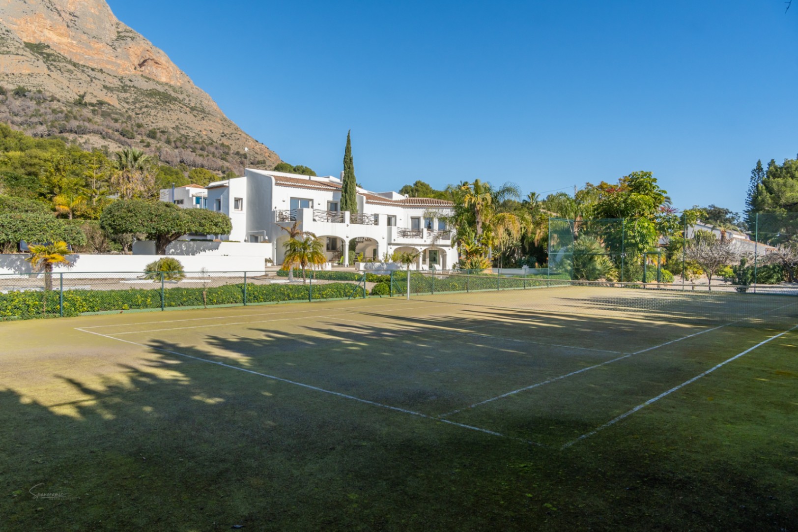 Privé-tennisbaan met groen oppervlak en omheining, gelegen voor de witte villa in Ibiza-stijl met het Montgó-gebergte op de achtergrond.