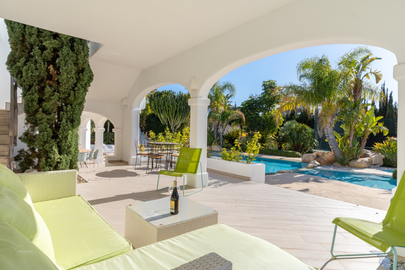 Schaduwrijke naya met witte bogen en limoenkleurig meubilair, uitkijkend op het zwembad en de volgroeide mediterrane tuin met palmbomen.