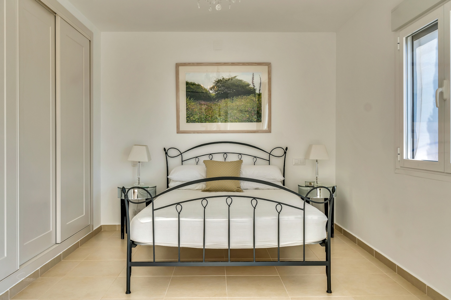 Slaapkamer met een zwart smeedijzeren bed, nachtkastjes van spiegelglas en crèmekleurige inbouwkasten. De ruimte is afgewerkt met neutrale tegels en zijdelingse lichtinval.