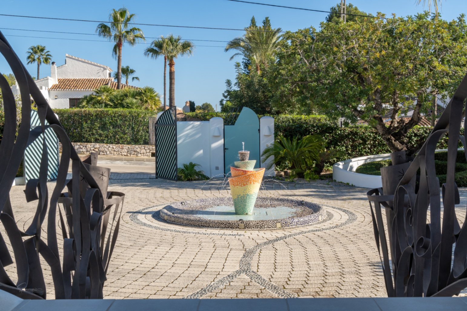 Uitzicht vanaf de entree met decoratief ijzerwerk op een geplaveid voorplein met een opvallende mozaïekfontein en volgroeide mediterrane beplanting.