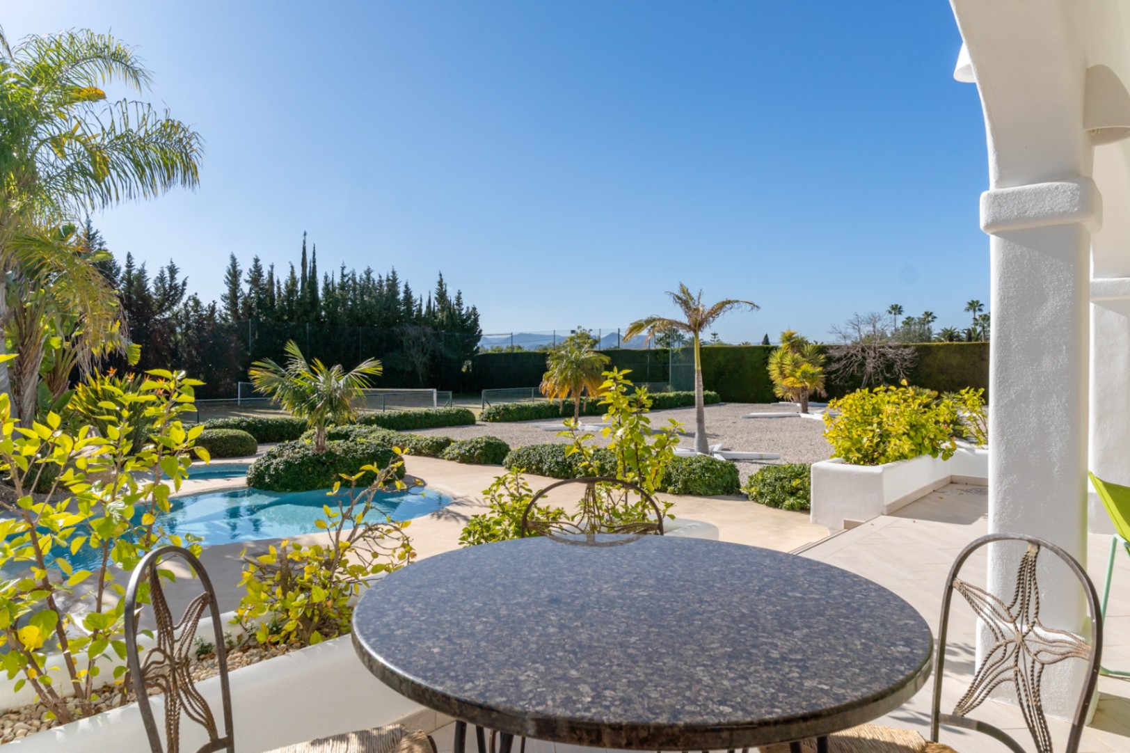Uitzicht vanaf de naya met granieten tafel op het zwembad, de aangelegde mediterrane tuin en de privétennisbaan.