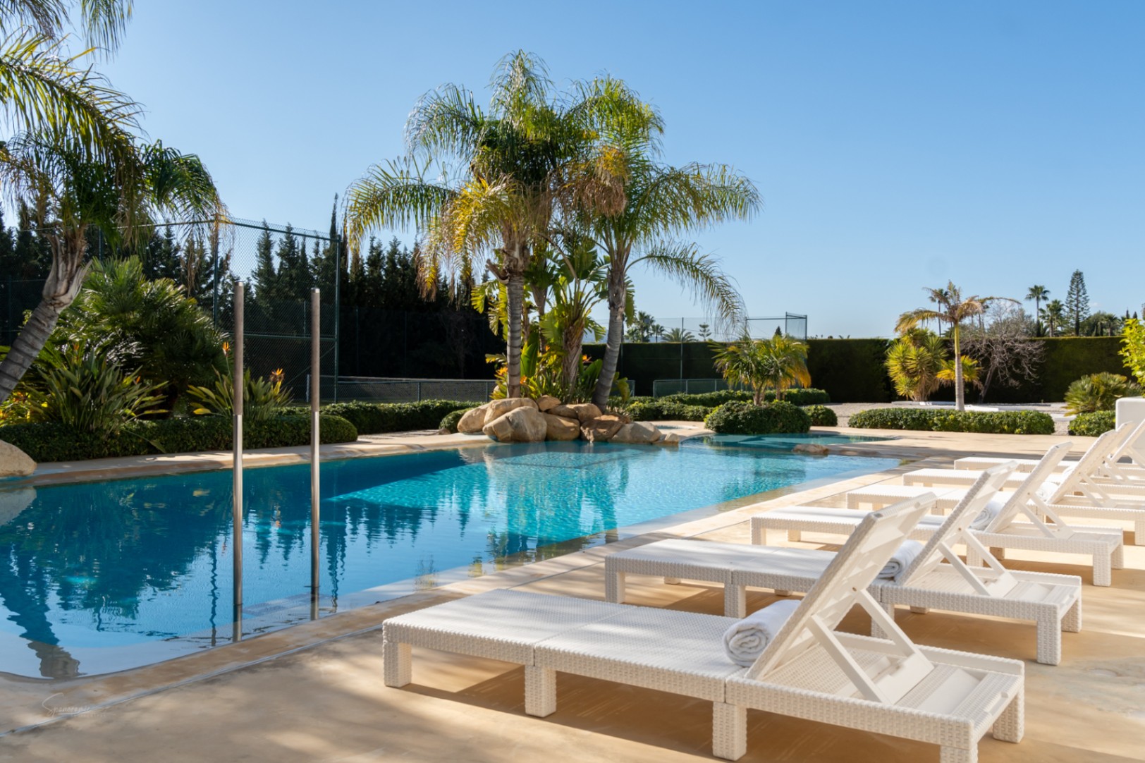 Zwembad van 70m² met witte rotan ligbedden, tropische palmbomen en natuurlijke rotspartijen, grenzend aan een eigen tennisbaan in een mediterrane tuin.