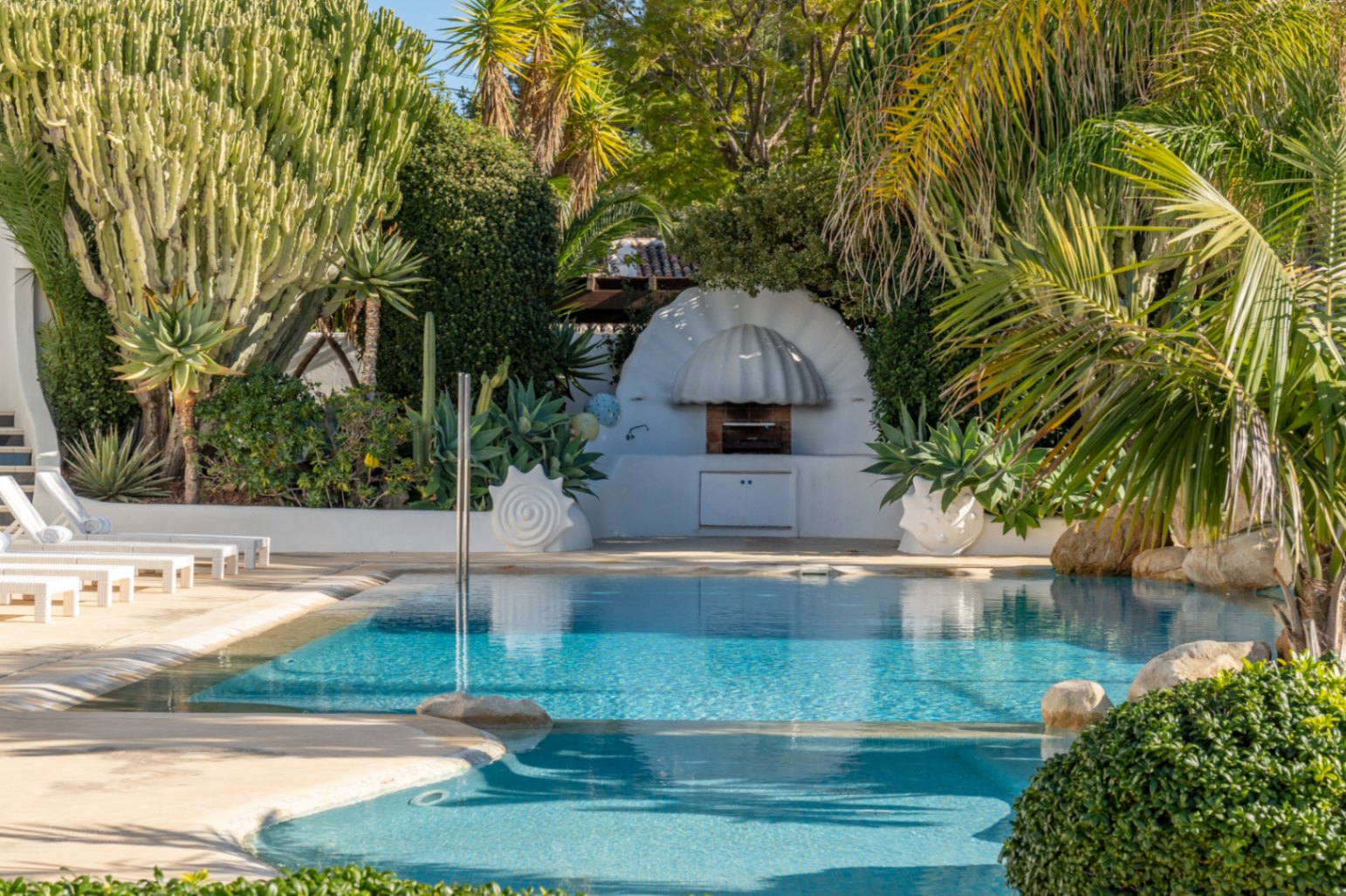 Zwembadgedeelte met een witte barbecue in organische stijl, omgeven door weelderig mediterraan groen, grote cactussen en witte ligbedden op een licht stenen terras.