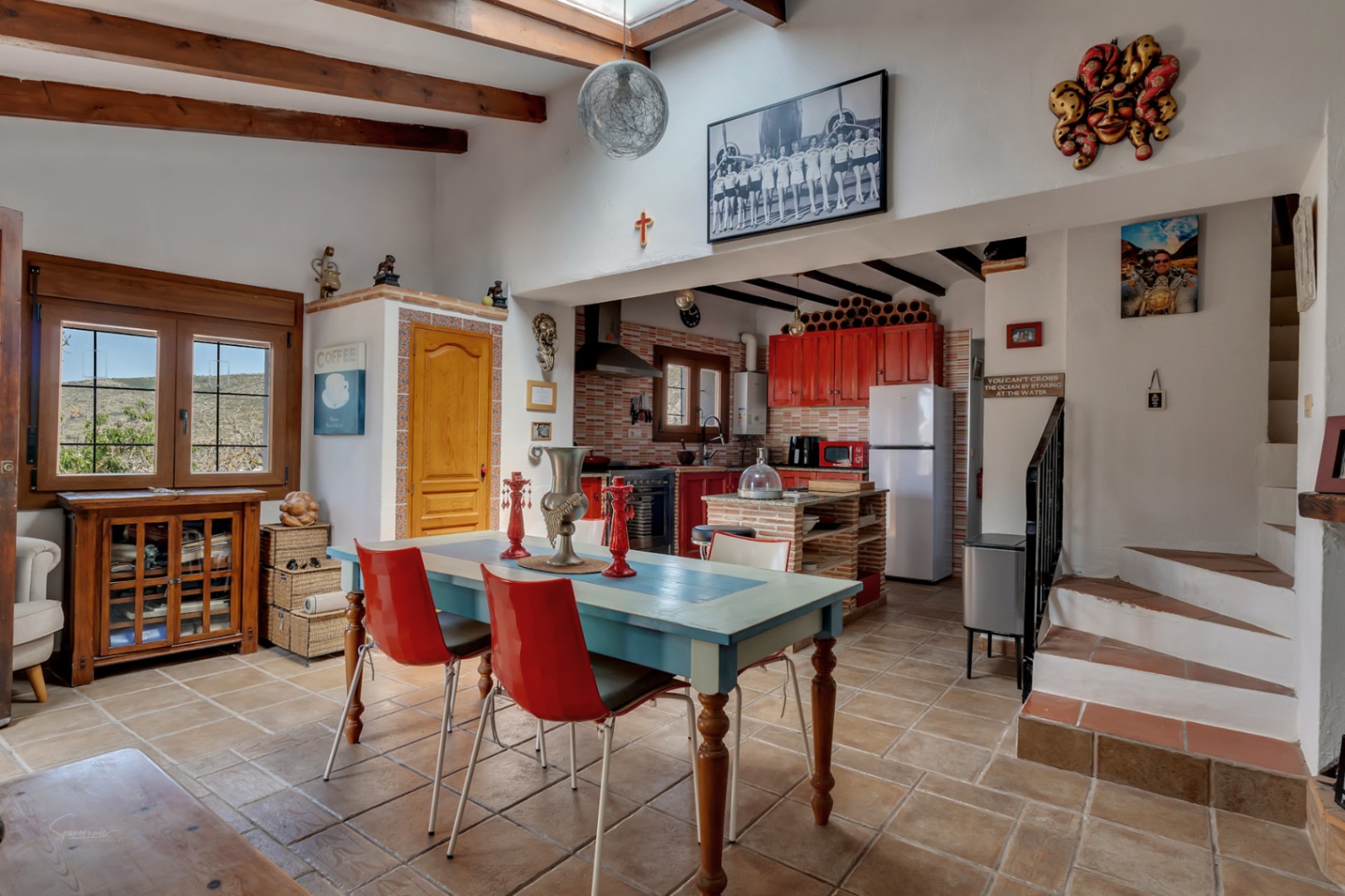Lichte eetkamer met blauwe tafel, rode stoelen en rode keuken. Houten balken en eclectisch decor.