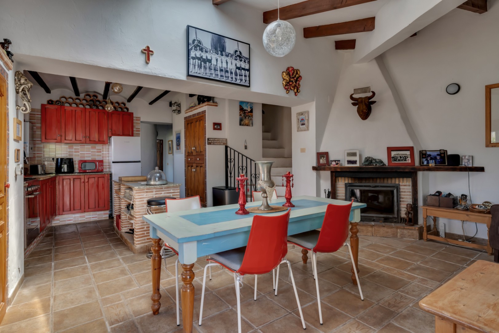 Rustieke eethoek met blauwe tafel, rode stoelen en een rode houten keuken. Open haard met decor en trap.