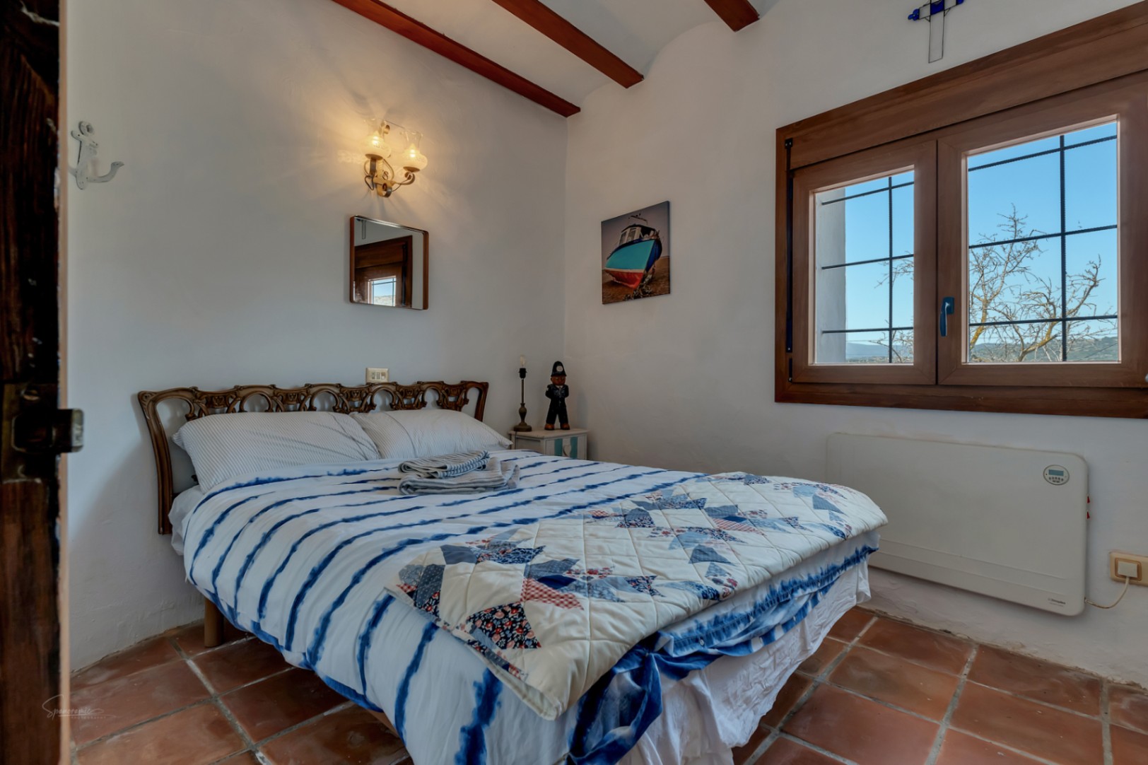Rustieke slaapkamer met terracotta vloer, een bed met een blauw quilt en een houten raam met uitzicht naar buiten.