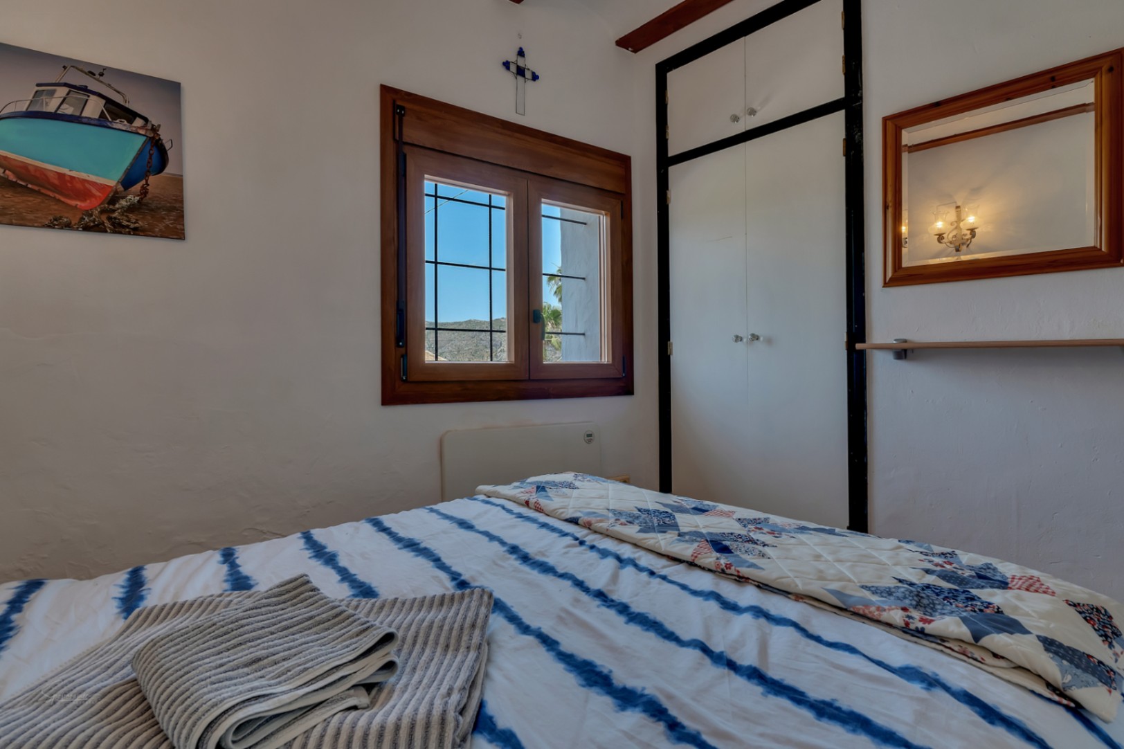 Slaapkamer met houten raam, witte kast en blauw gestreept beddengoed. Rustieke inrichting en heldere sfeer.