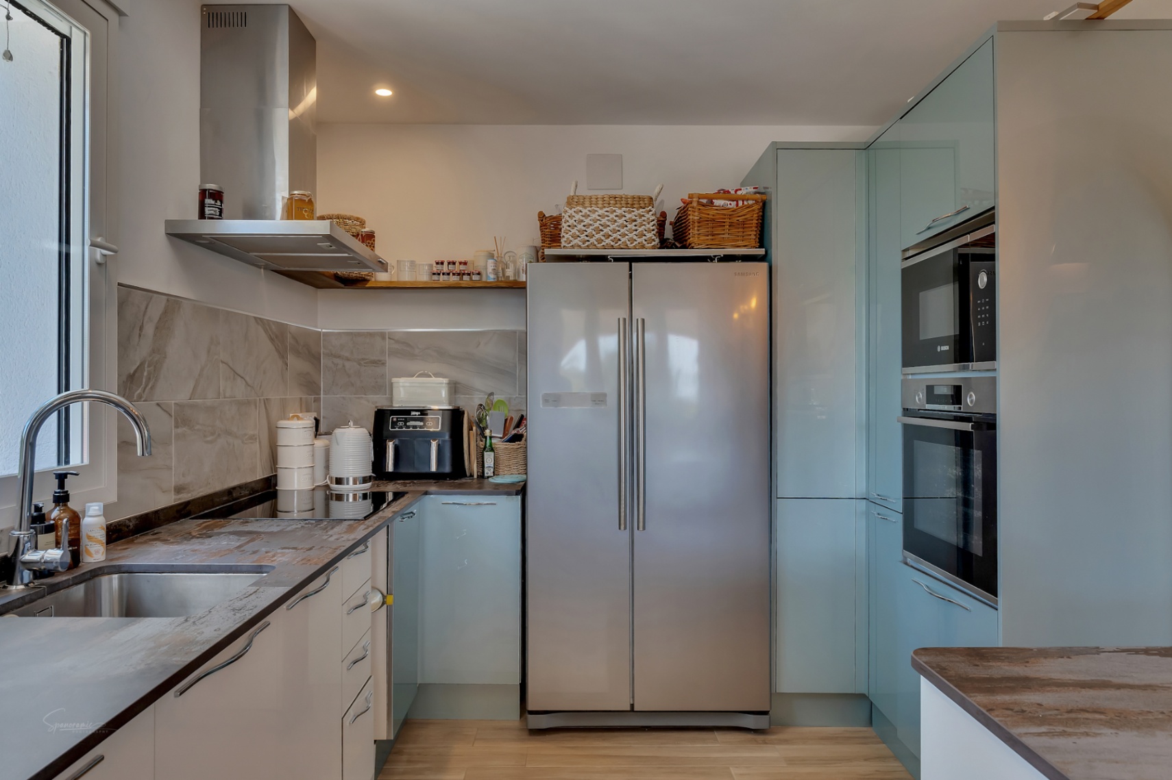 Keuken met stenen werkbladen, roestvrijstalen apparaten en lichtblauwe kasten.