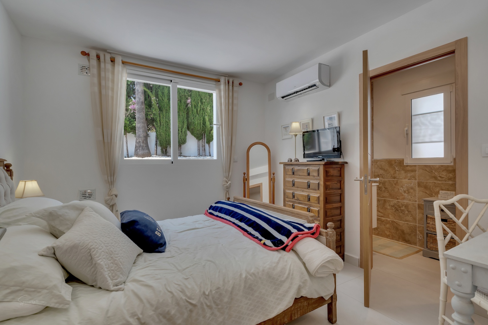 Lichte slaapkamer met houten bedframe, rustieke ladekast en en-suite badkamer met aardetinten tegels.
