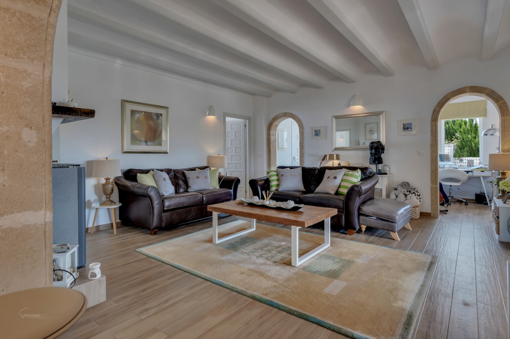 Woonkamer met leren banken, een vloerkleed met print en witte balken aan het plafond. Stenen bogen verbinden met andere kamers.