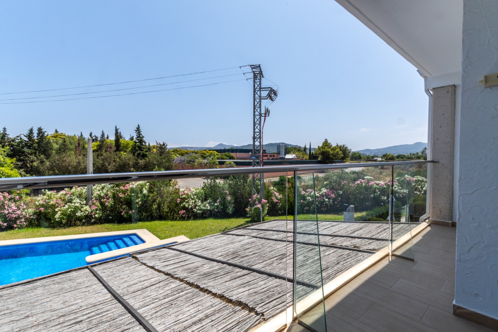Bovenliggend terras met glazen balustrade en uitzicht op het privépatioterras met rieten overkapping, het zwembad en de heuvels van Jávea.