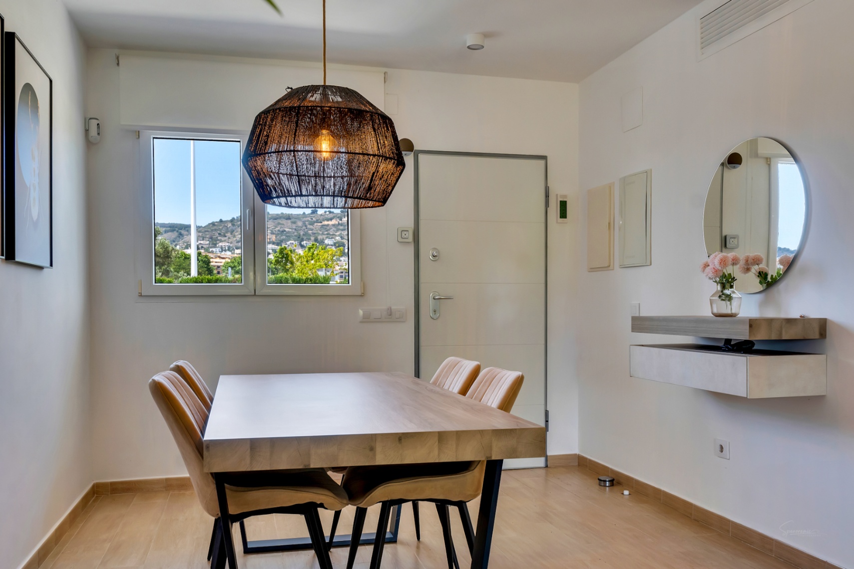 Eethoek met een massief houten tafel, gestoffeerde stoelen en een geweven hanglamp. Een zwevend wandmeubel met ronde spiegel siert de lichte ruimte met uitzicht op de heuvels.