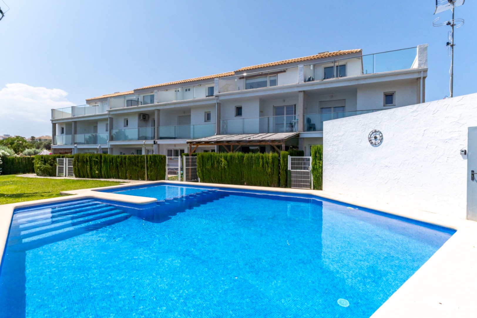Gemeenschappelijk zwembad met blauwe mozaïektegels voor de moderne witte gevel van de woningen, voorzien van glazen balustrades en privétuinen met hagen.