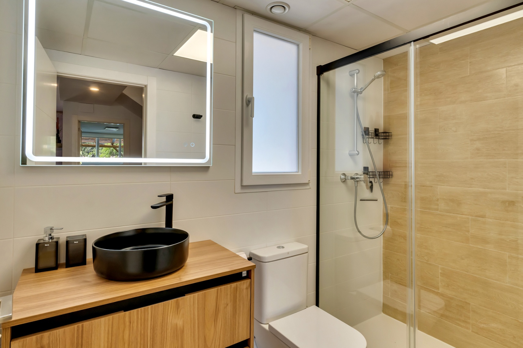 Moderne badkamer met zwarte opzetwastafel op een houten meubel, verlichte spiegel en inloopdouche met zwart profiel en tegels met houteffect.