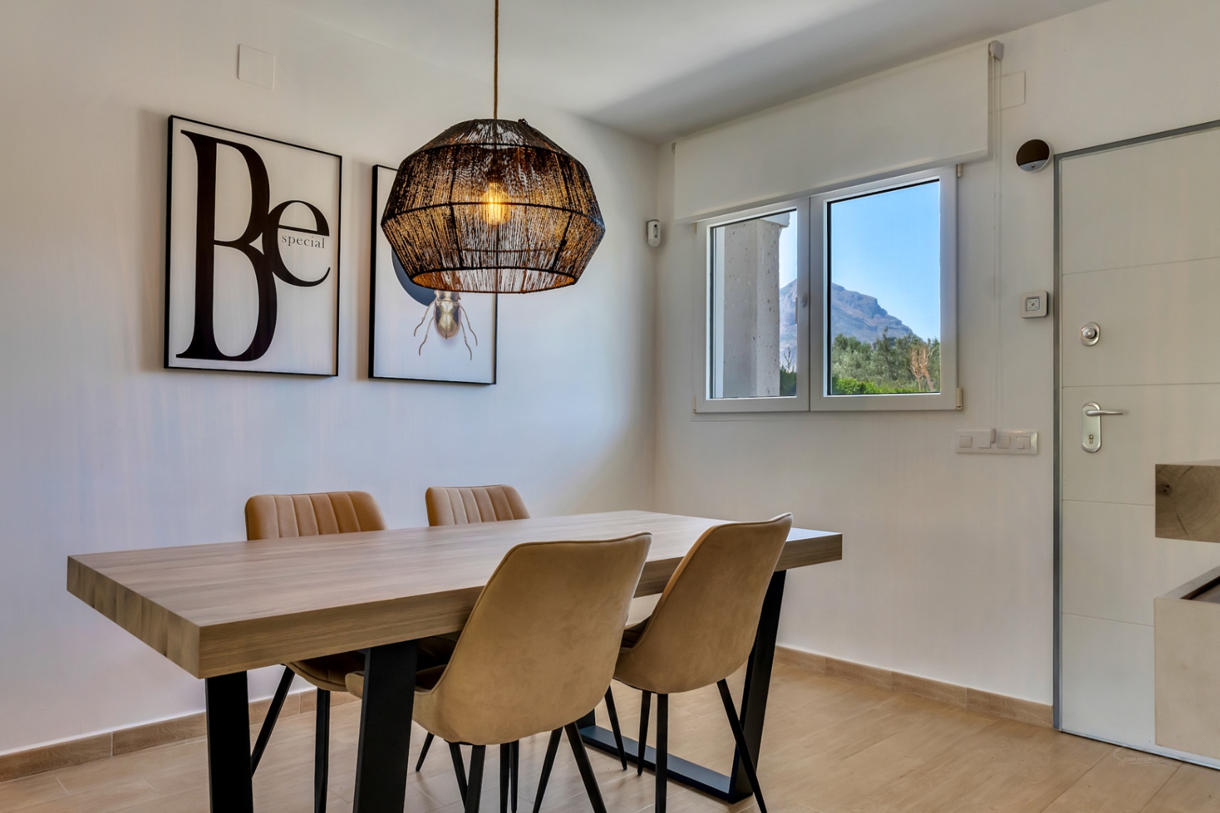 Moderne eethoek met houten tafel, gestoffeerde stoelen en een geweven hanglamp. Het raam biedt uitzicht op de iconische Montgó-berg.