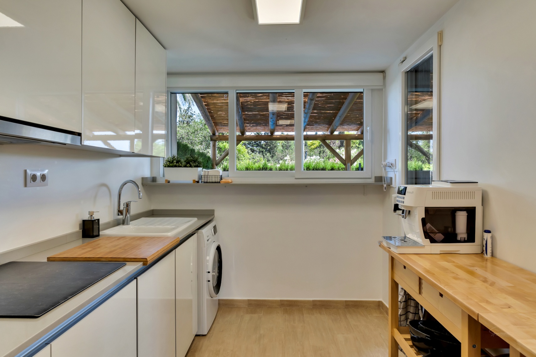 Moderne keuken met hoogglans witte kasten, geïntegreerde wasruimte en een groot raam met uitzicht op de houten pergola van de privépatiotuin.