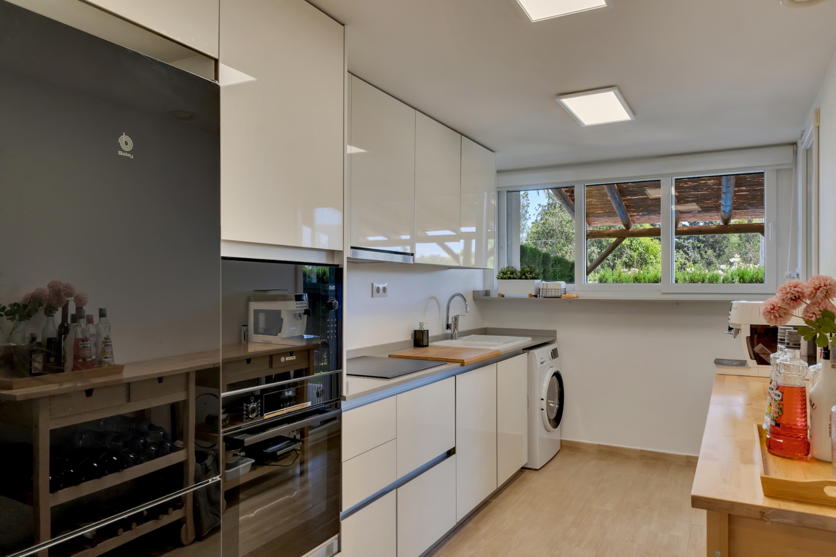 Moderne keuken met witte hoogglans kasten, houten werkbladen en inbouwapparatuur. Een groot raam zorgt voor natuurlijk licht en uitzicht op de privépatiio.