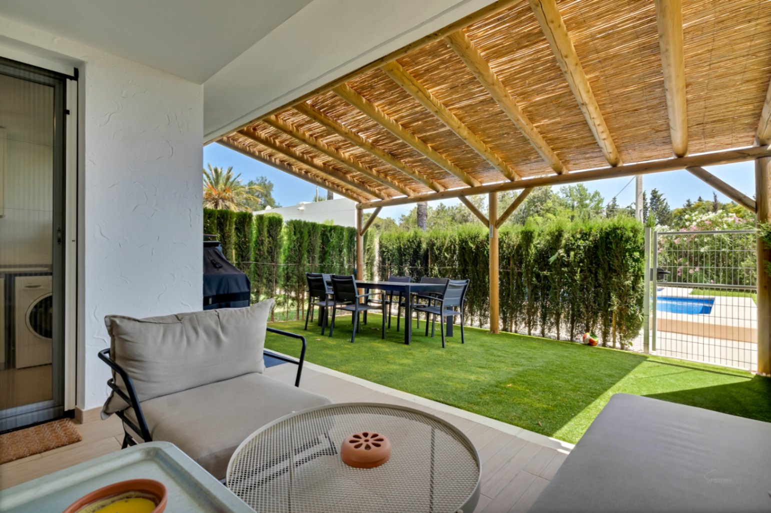 Privépatio met houten pergola en rieten dak, buiten-eethoek en kunstgras. Een poortje biedt directe toegang tot het gemeenschappelijke zwembad en de tuin.