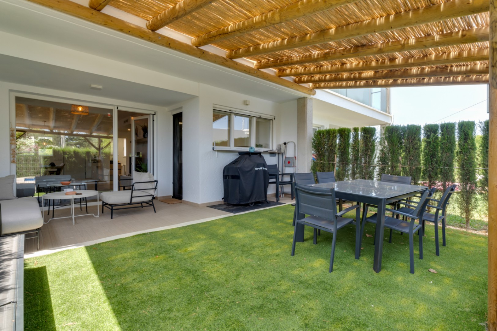 Privépatio met houten pergola en rieten zonwering, eethoek buiten en kunstgras, verbonden met de woonkamer via grote glazen schuifdeuren.