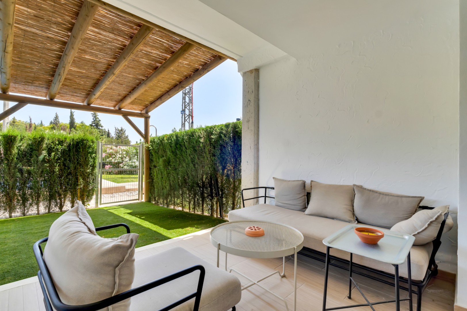 Privépatio met zithoek onder een houten pergola, kunstgras en een metalen poort die directe toegang biedt tot de gemeenschappelijke ruimtes.