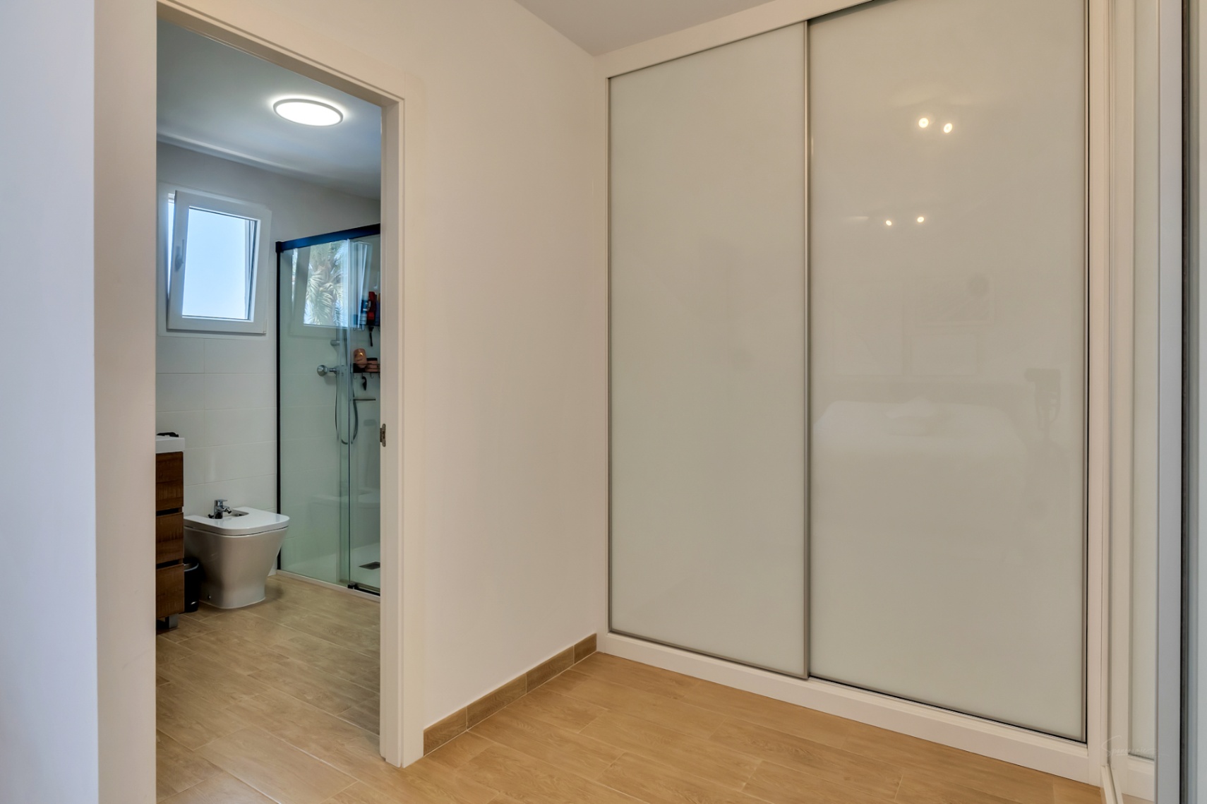 Slaapkamer met grote inbouwkast met witte schuifdeuren en directe toegang tot de aangrenzende moderne badkamer met inloopdouche.