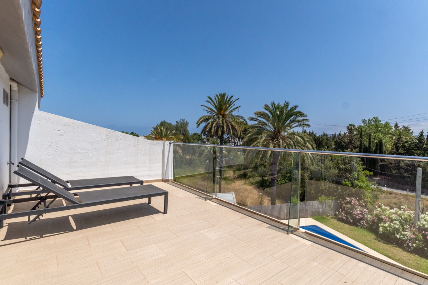 Terras op de bovenverdieping met keramische tegels, glazen balustrade en ligbedden, uitkijkend over palmbomen en de groene omgeving.