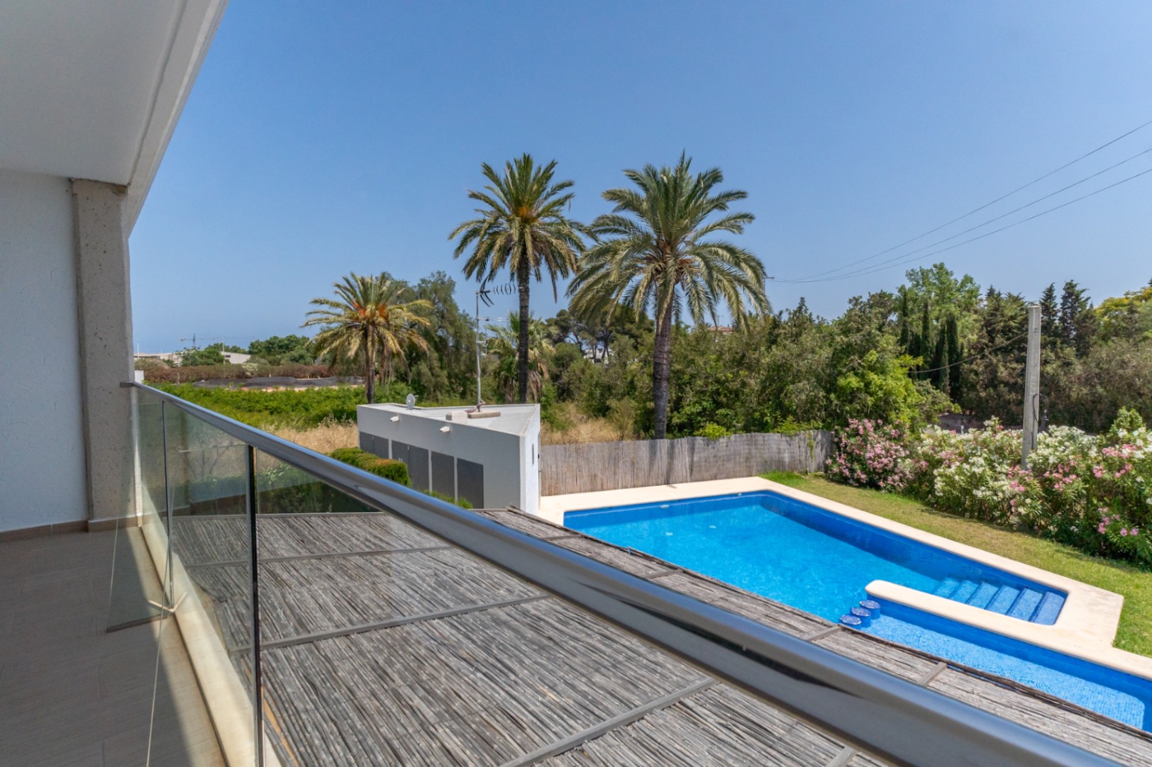 Uitzicht vanaf het boventerras met glazen balustrade op het gemeenschappelijke zwembad, de mediterrane tuin met palmbomen en de houten pergola van de patio.