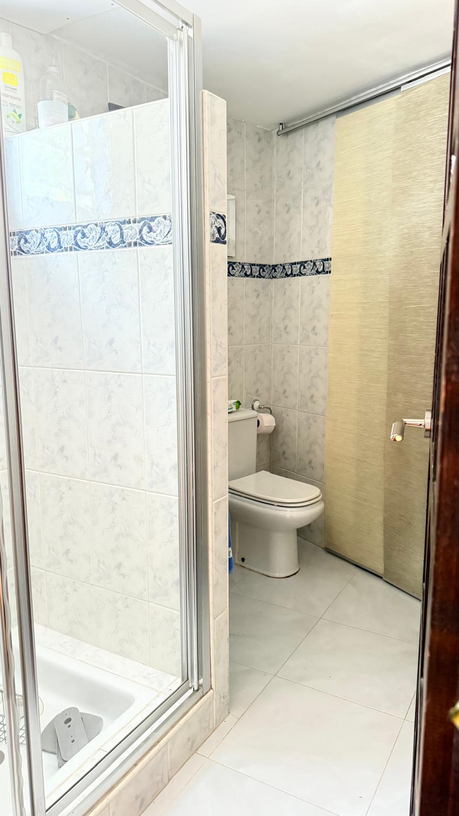 Badkamer met glazen douchecabine, witte wandtegels met een blauwe sierrand en een wit toilet. De ruimte is voorzien van een getextureerde schuifdeur.