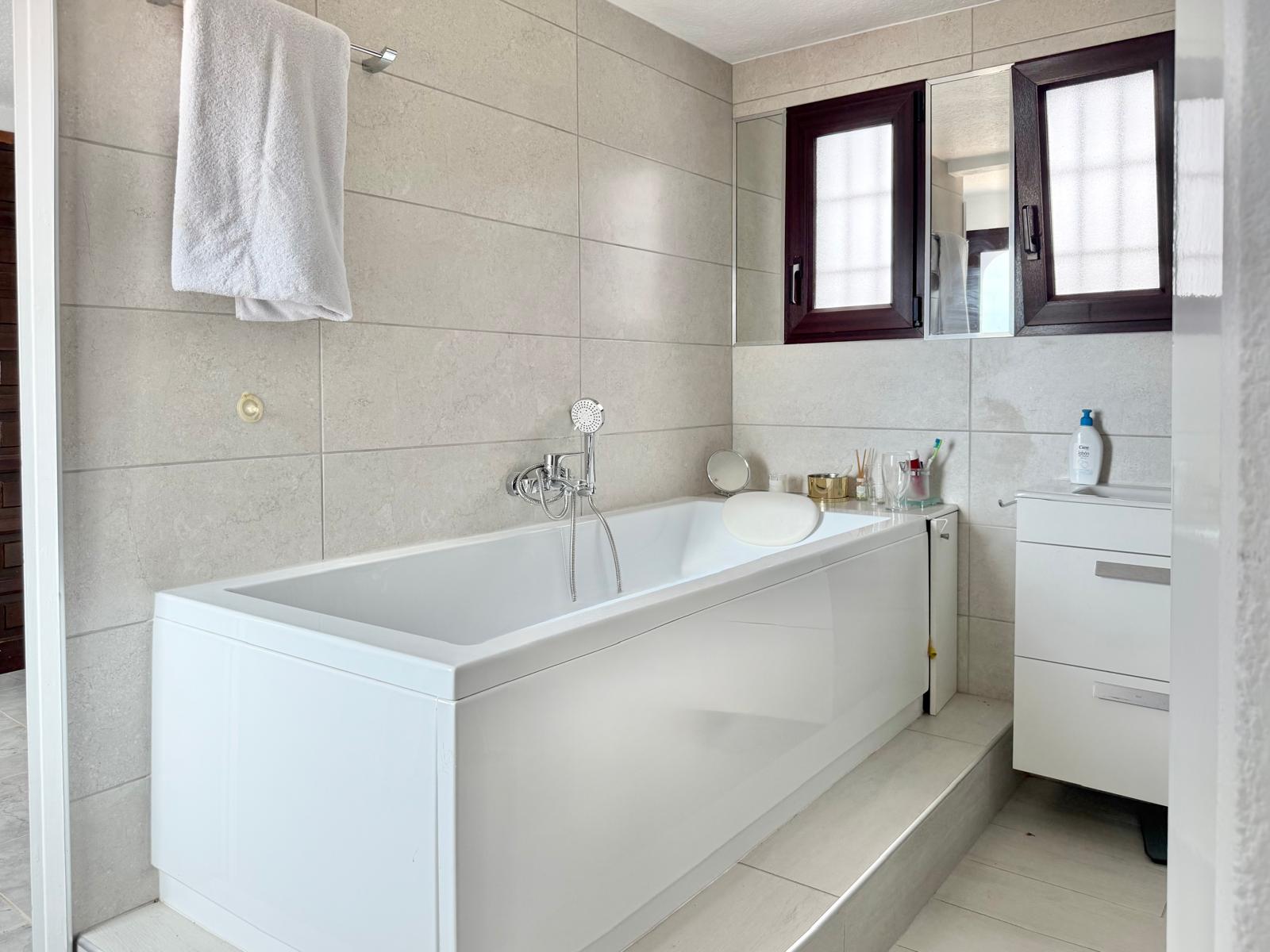 Badkamer met wit ligbad en hoofdsteun, grootformaat keramische wandtegels in neutrale tinten en een wit wastafelmeubel naast ramen met donkere kozijnen.