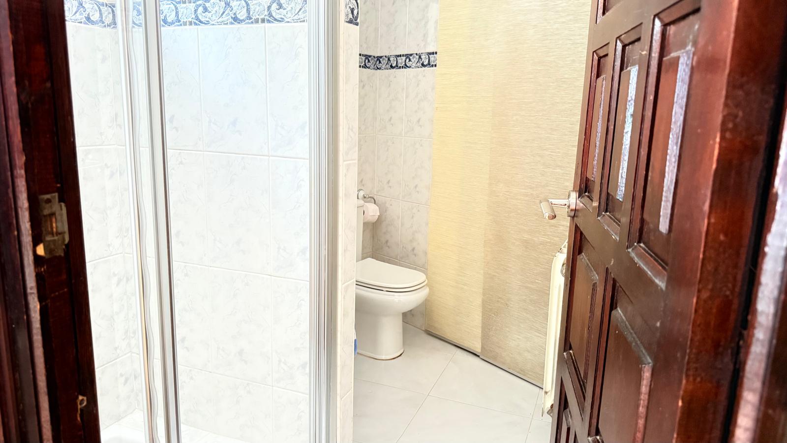 Badkamer met witte wandtegels en een blauwe sierrand, voorzien van een wit toilet en een glazen douchecabine, toegankelijk via een donkerhouten paneeldeur.