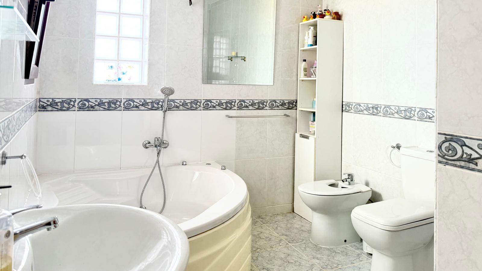 Badkamer voorzien van een groot hoek-hydromassagebad, bidet en toilet, afgewerkt met witte wandtegels en een klassieke decoratieve sierrand.