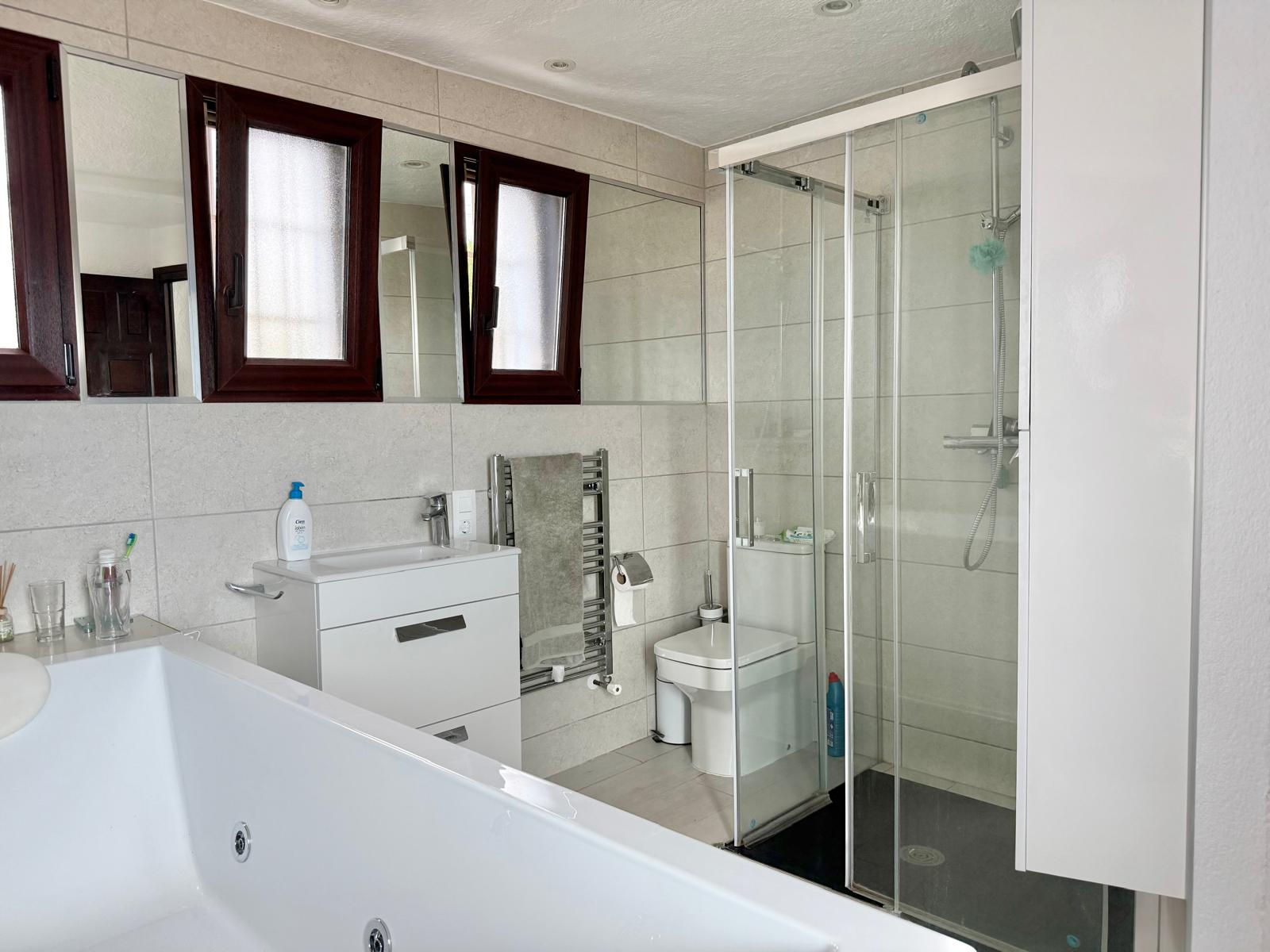 Badkamer voorzien van een hydromassagebad, een aparte glazen douchecabine, een wit wastafelmeubel en een handdoekradiator tegen een neutraal betegelde wand.