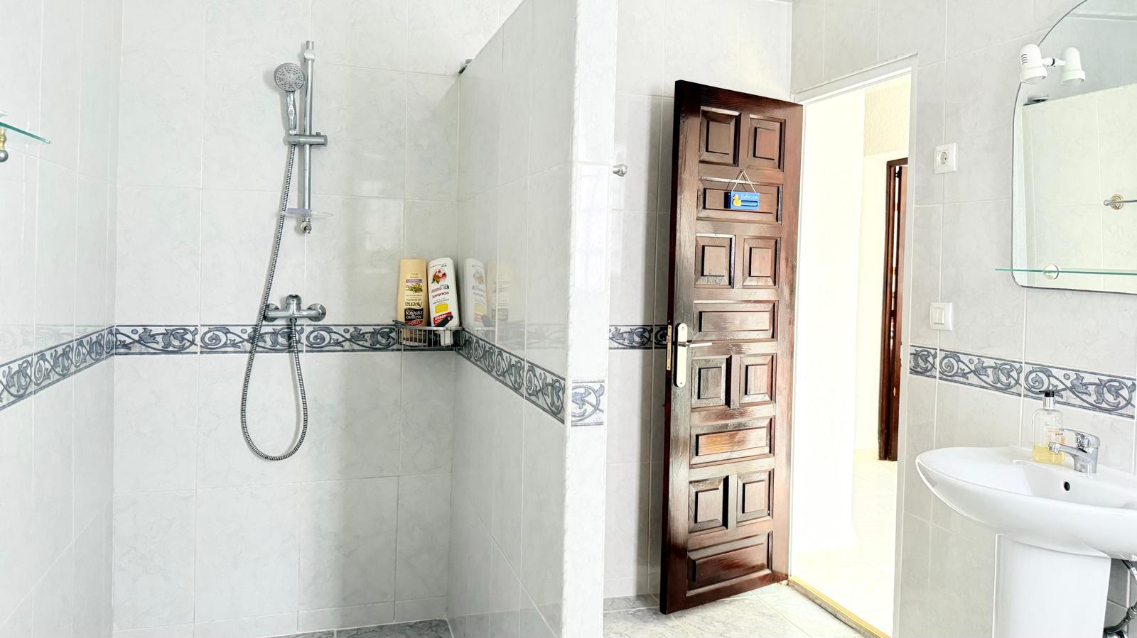 Badkamer voorzien van een inloopdouche met decoratieve tegelrand, een kolomwastafel en een massief houten paneeldeur.