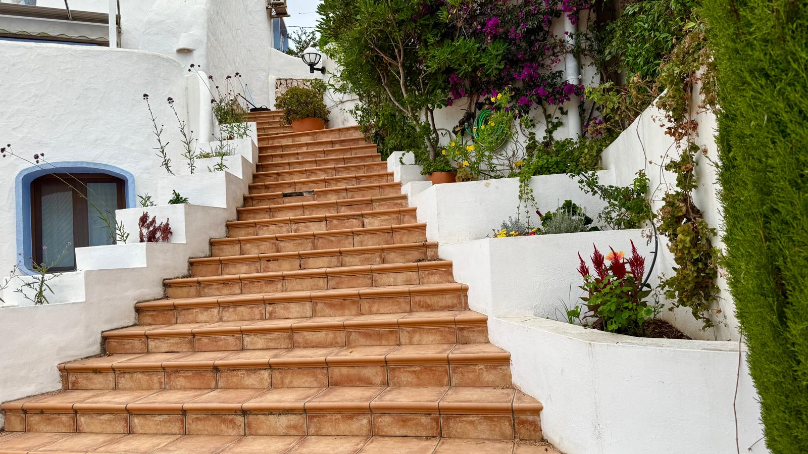 Buitentrap met terracotta tegels die de verschillende niveaus van de villa verbindt, geflankeerd door witte plantenbakken met mediterrane beplanting en bougainville.