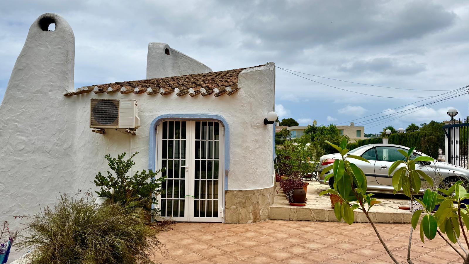Exterieur in Ibiza-stijl met ronde witte muren, blauw omrande glazen deuren en een betegeld terras grenzend aan de parkeergelegenheid.