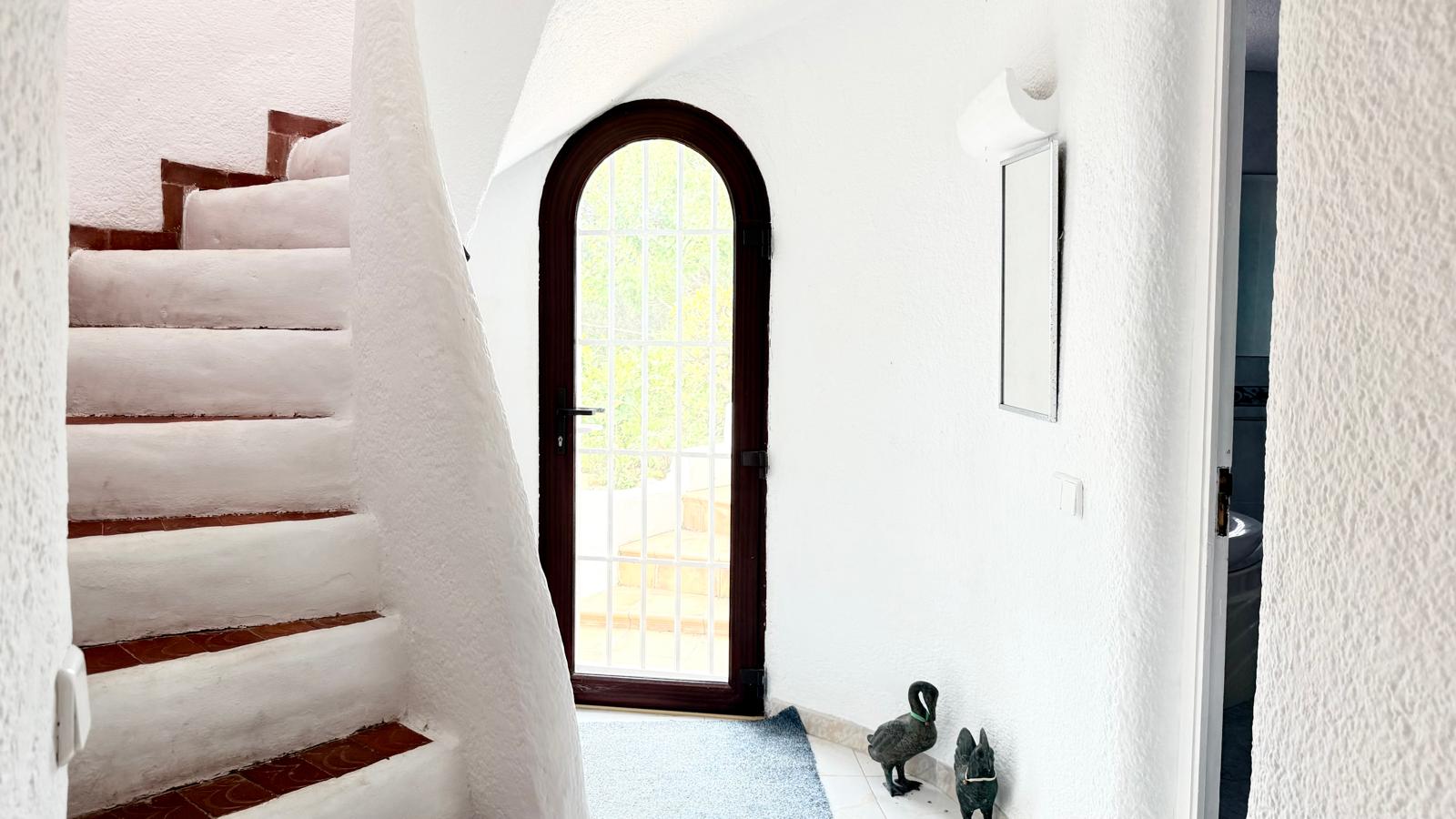 Gebogen binnentrap in Ibiza-stijl met terracotta treden en witte structuurwanden, gelegen naast een karakteristieke boogdeur met toegang tot buiten.