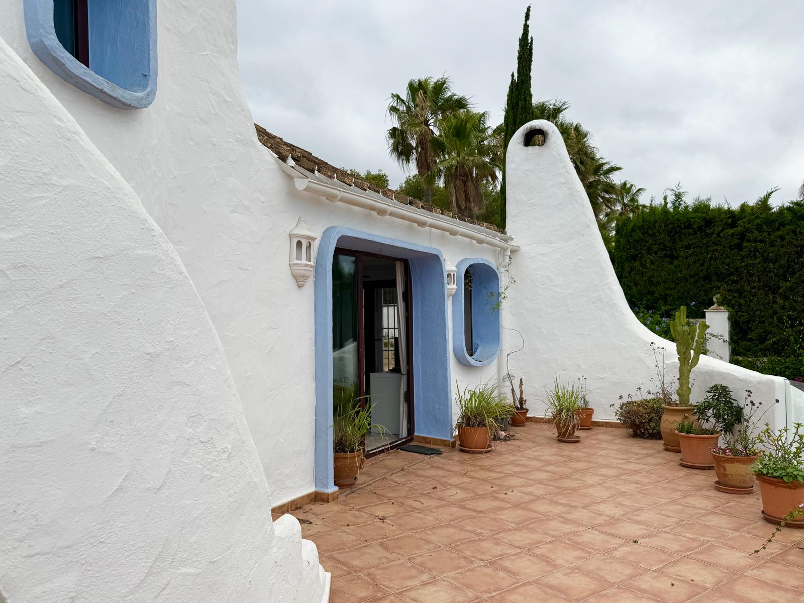 Gevel in Ibiza-stijl met afgeronde witgekalkte muren, blauwe kozijnen en een terracotta terras met mediterrane planten en een karakteristieke schoorsteen.