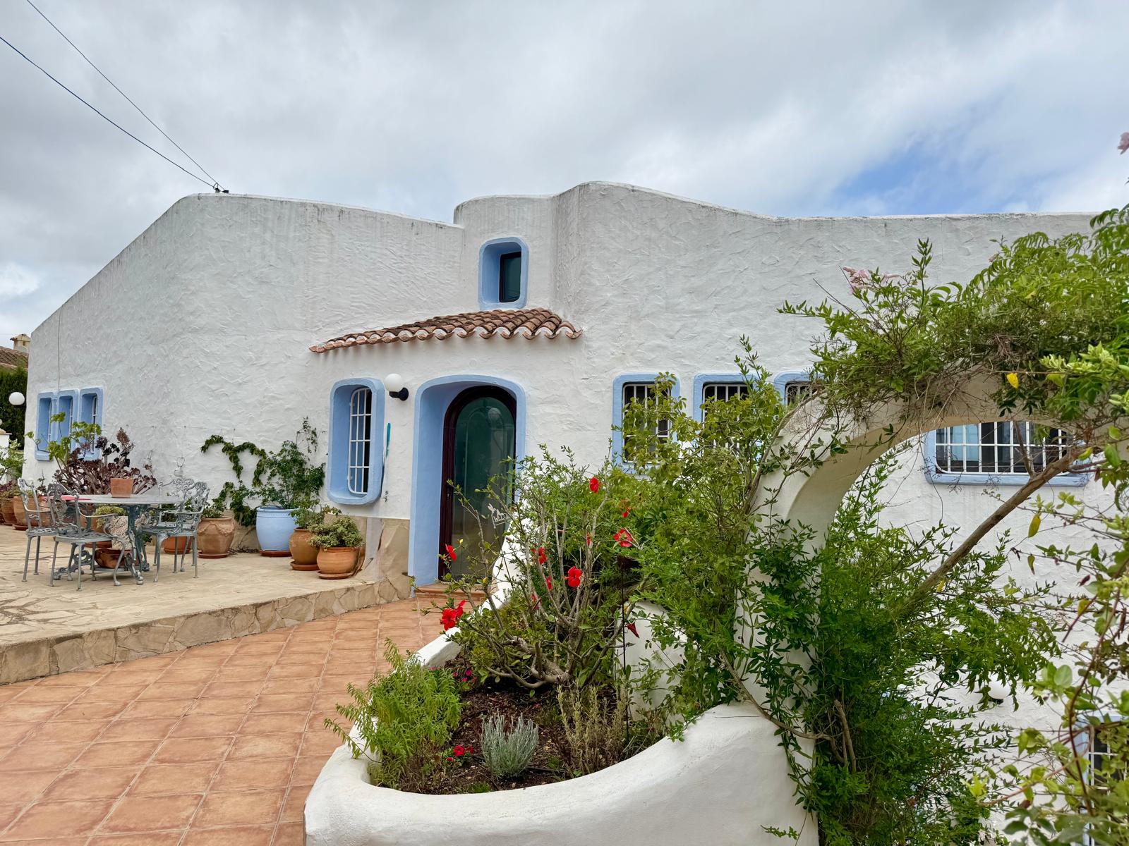 Gevel in Ibiza-stijl met organische witte muren, blauwe kozijnen en een terracotta terras omgeven door mediterrane planten en decoratieve potten.
