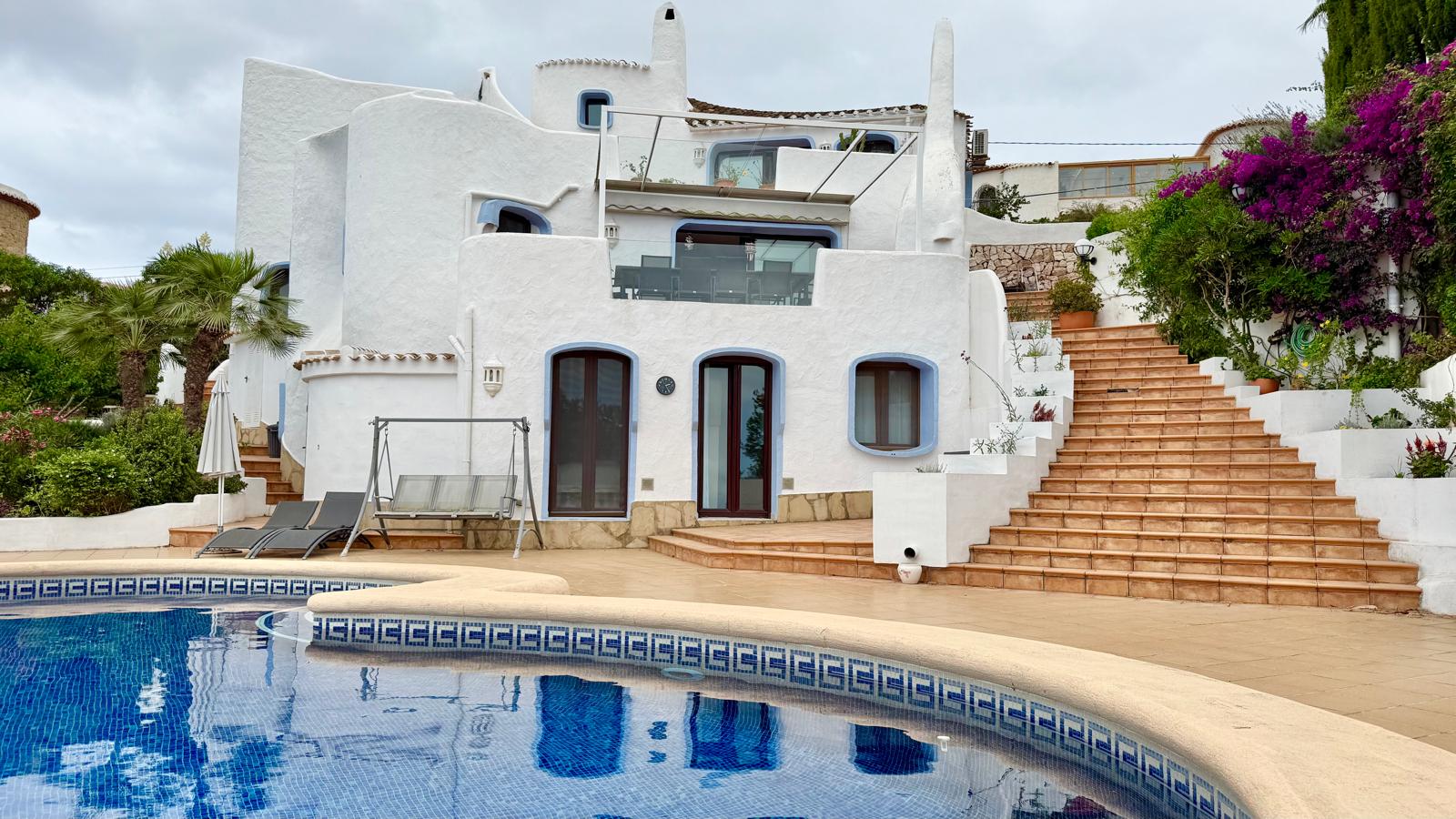 Gevel in Ibiza-stijl met organische witte vormen en terrasvormige niveaus, verbonden door buitentrappen naast het zwembad en de tuin.