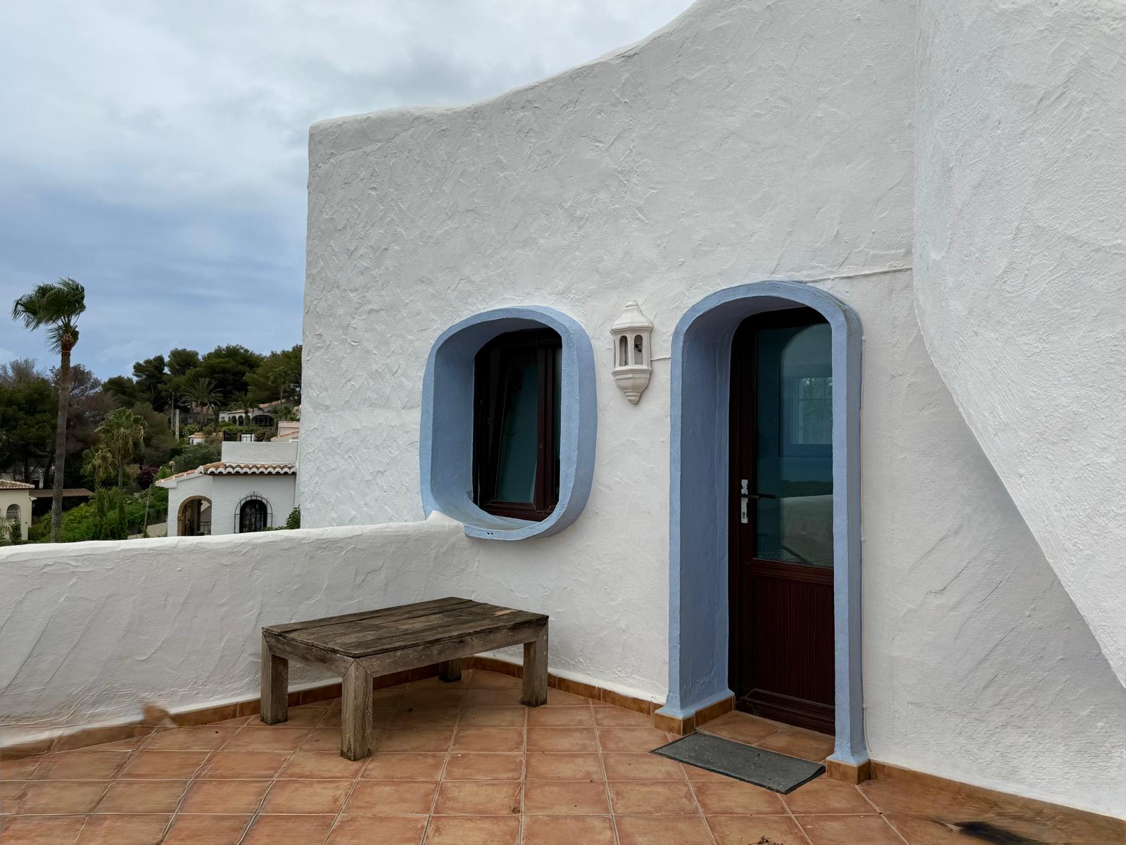 Gevel in Ibiza-stijl met witte muren met textuur, blauwe boogkozijnen en terracotta tegels. Het terras is voorzien van donkerhouten schrijnwerk en rustiek meubilair.