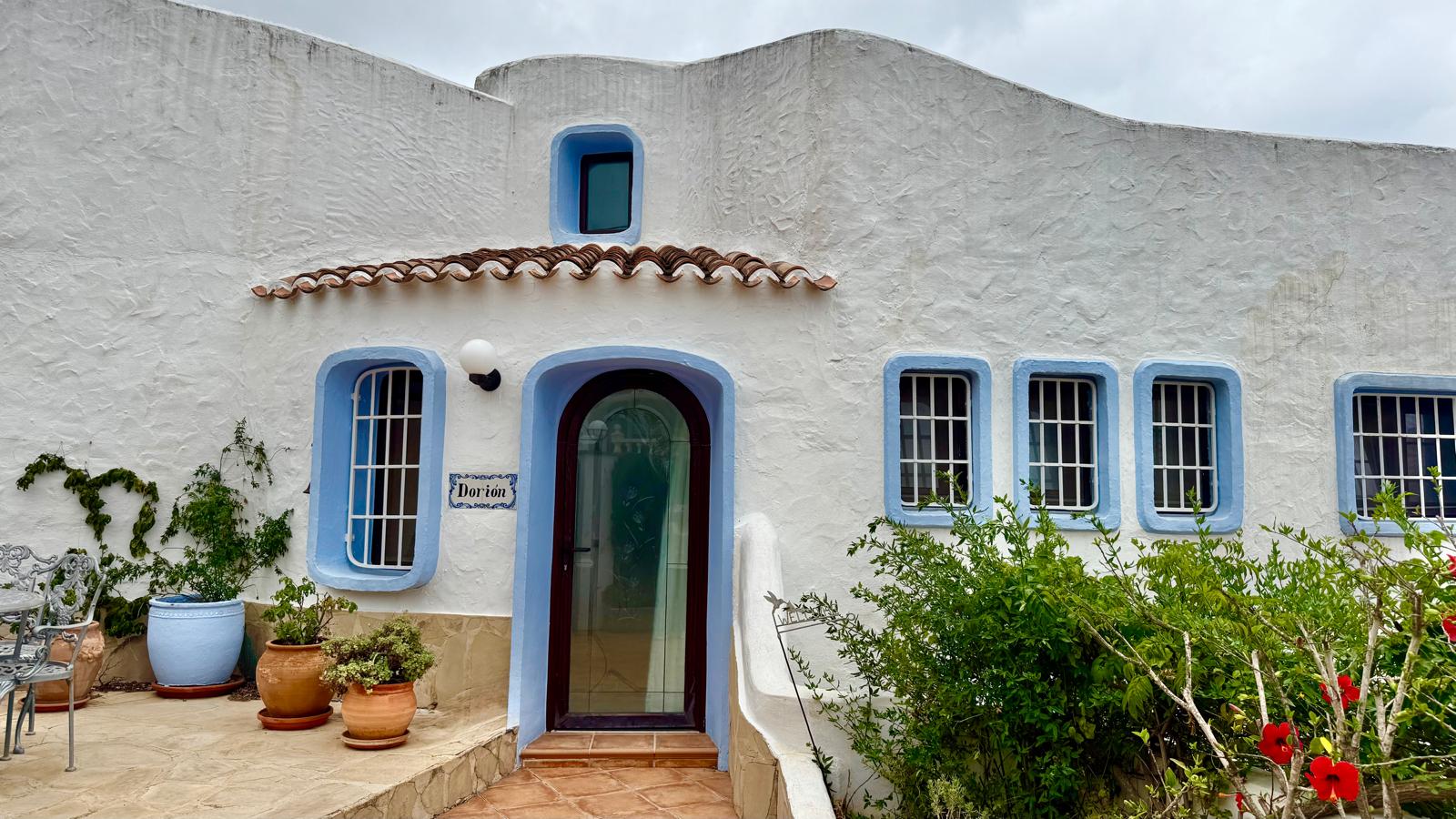 Gevel in Ibiza-stijl met witte structuurmuren, blauwe raamkozijnen en een boogdeur. De entree is afgewerkt met terracotta tegels en mediterrane beplanting.