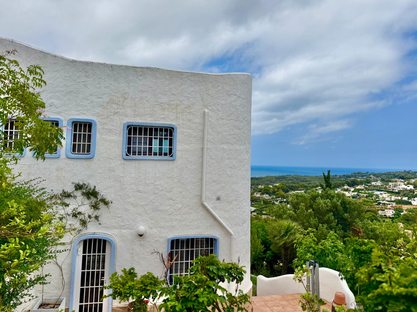 Gevel in Ibiza-stijl met witte structuurmuren, blauwe raamkozijnen met tralies en een panoramisch uitzicht over de Middellandse Zee en de kustlijn van Jávea.