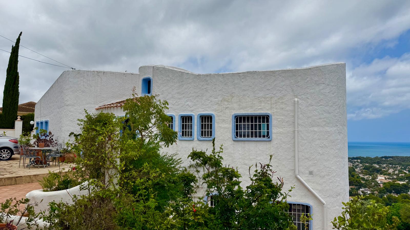Gevel in Ibizastijl met witte getextureerde muren, blauwe kozijnen en organische lijnen, gelegen op een verhoogde positie met panoramisch uitzicht op zee.
