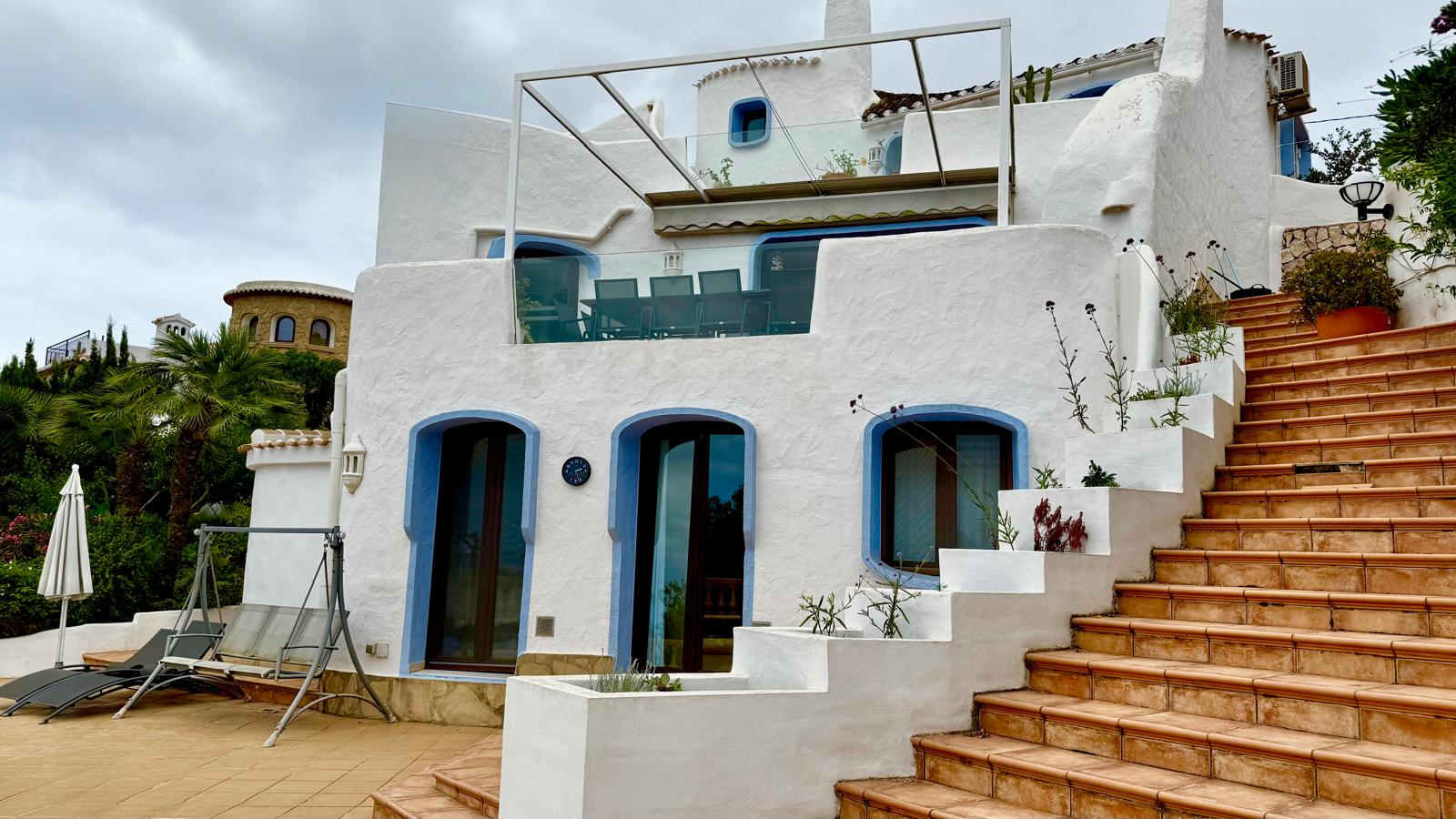Gevel in Ibizastijl met witte muren en blauwe kozijnen. De verschillende niveaus zijn verbonden via terracotta trappen en een terras met glazen balustrade en pergola.
